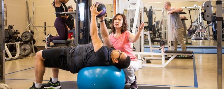 An exercise coach working with a student who is lifting weights on an exercise ball.